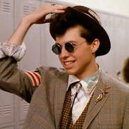 a picture of duckie from pretty in pink. he is wearing a pork pie hat and circular sunglasses. he is fixing his hat and smiling